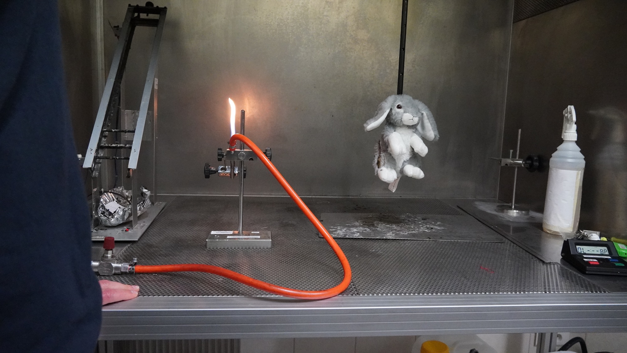 Metalowy boks stanowiska do sprawdzania łatwopalności zabawek. Zapalony palnik Bunsena obok pluszaka zawieszonego na metalowym uchwycie.